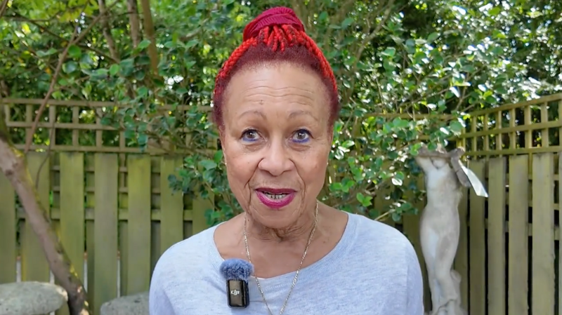 A woman with red hair and a grey t-shirt stands in a garden in the sun while speaking to the camera