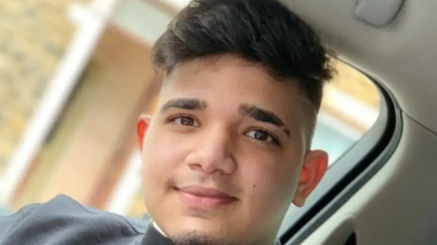 A young man seated inside a vehicle. He has short, dark hair with the sides cut shorter than the top and is in the center of the frame looking towards the camera. He has light facial hair on his chin.