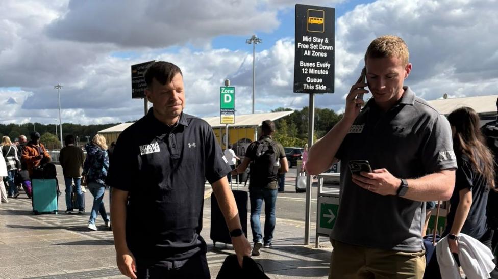 Two men standing outside an airport while other passengers walk around behind them