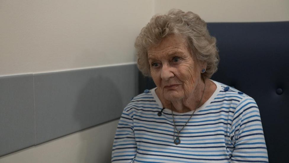 A screengrab of an older woman with shoulder-length wavy hair, wearing a blue striped top and sitting in a hospital corridor. 