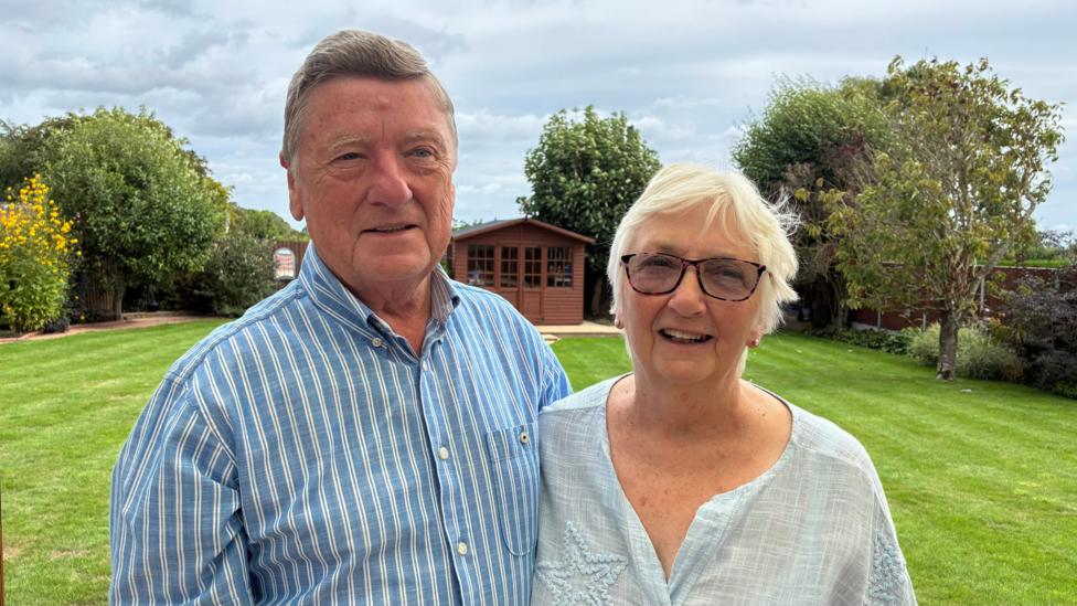 John Craggs, with brown hair and a blue and white striped shirt, stands next to his wife Libby, who is wearing a white top, and has white hair and glasses. In the background is their garden, with a neatly mowed green lawn and a brown garden shed.