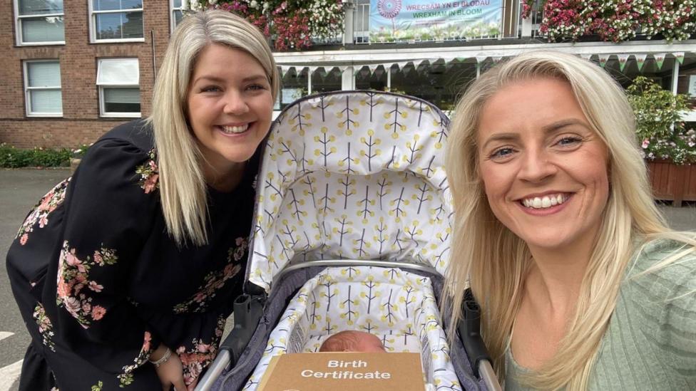 In the middle of the photo is a pram. In the bassinet is a baby, you can't see his face but you can see the side of face. The outside of the pram is grey and the inside is white, there are grey trees with yellow balls coming off of them on the inside. On top of the bassinet is a brown envelope with 'Birth Certificate' written on it the top in white writing. There are two women, one sitting either side of the pram. The woman on the left is bending down next to the pram, wearing a black dress with pink and red flowers on it, her blonde hair is down and she is smiling. The woman on the right is crouching next to the pram, taking the photo. She is wearing a green top, her blonde hair is down and she smiling.