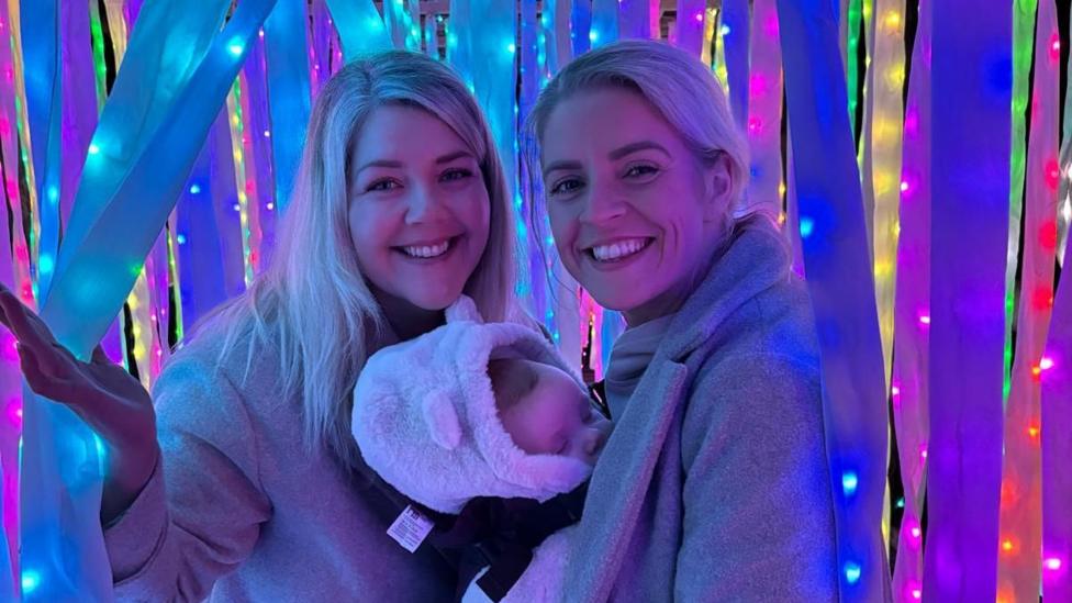 Two smiling, blonde, white women, wearing fluffy coats, standing amongst different coloured falling ribbons and fairy lights. A baby, dressed in a fluffy romper, is in a baby carried on the front of one of the women while he sleeps. There is a deep blue tinge to the photo.