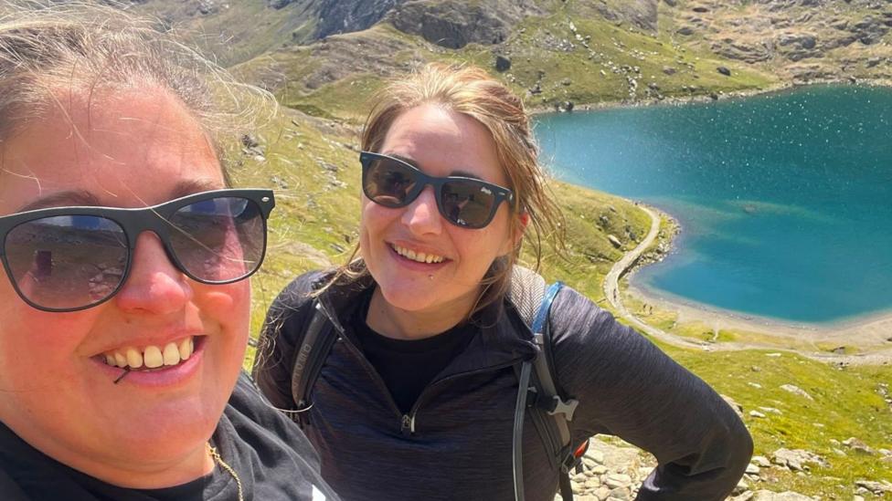 Two white women in sunglasses smiling on a mountain side. There's a blue lake in the background, and the terrain is grassy and rocky. Both are wearing black, their hair is tied up and they have small lip piercings. 