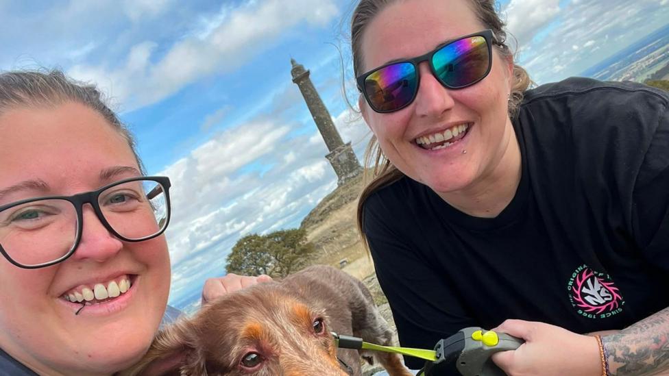 Two white women are smiling high up on top of a hill. One is wearing a black t shirt with a pink and white logo, and colouful sunglasses, you can see part of arm which has tattoos on it. She is holding a grey and yellow dog leg. Between the two women is a brown dachshund. The other woman is wearing clear glasses and is stroking the dog. Behind them is a monument on top of a mount. 