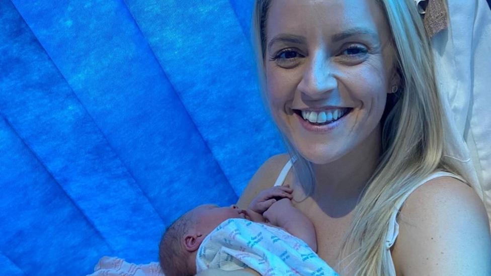 A white blonde woman in a white vest top smiling while holding a new-born baby in front of a blue hospital curtain. 