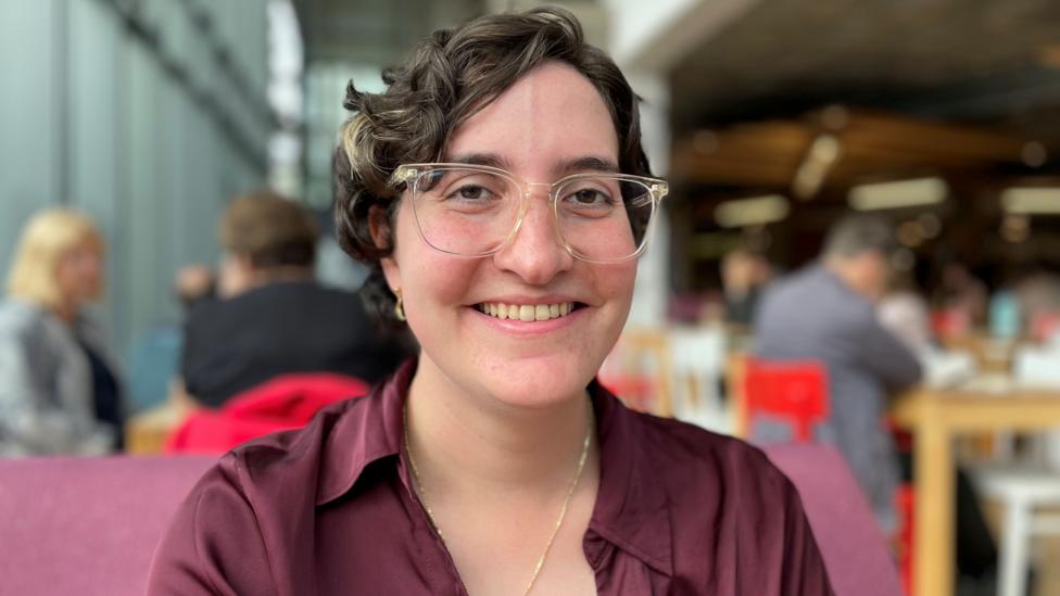 A woman with dark hair and glasses, wearing a maroon shirt, is smiling at the camera