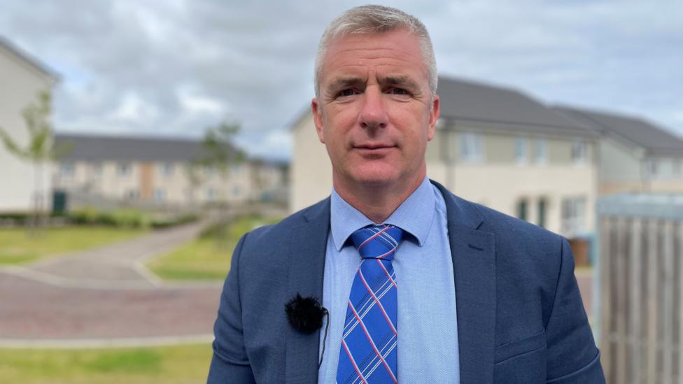 A headshot of a man with grey hair and wearing a blue suit, blue shirt and blue tie with houses in the background.