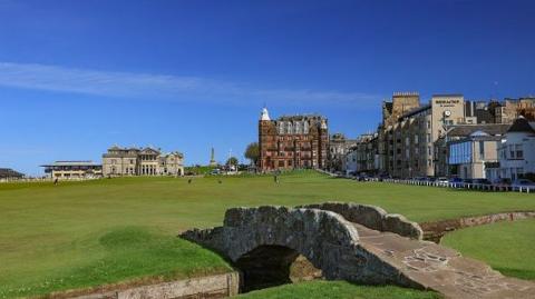 An old footbridge is in the foreground of a shot of a golf course with bright green grass, and a bright blue sky above