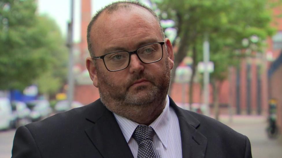A man with dark short hair and beard wearing glasses and a suit.
