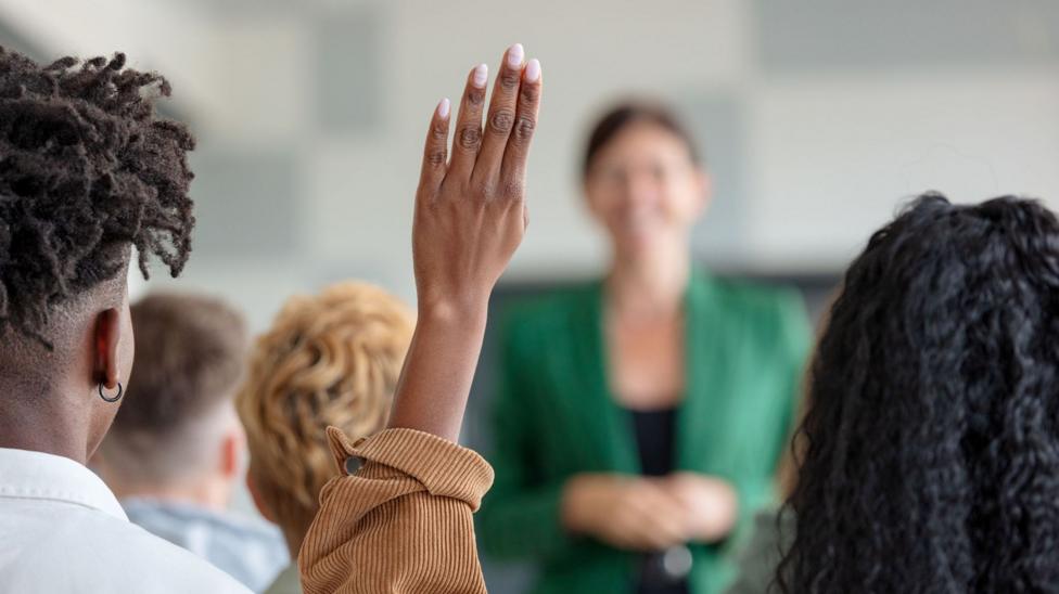 A participant raises a hand during a group training session while a lecturer stands at the front.