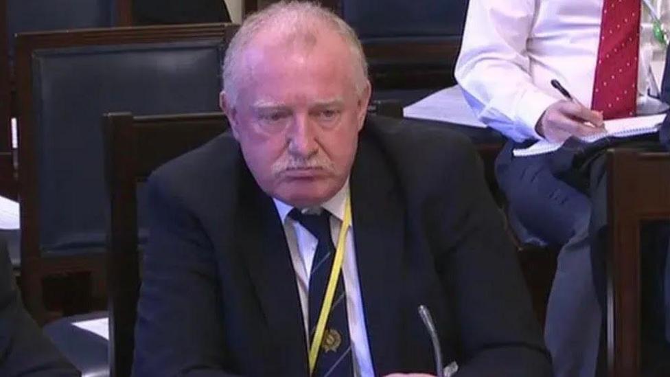 A man with grey hair, wearing a suit and tie with a yellow lanyard on.