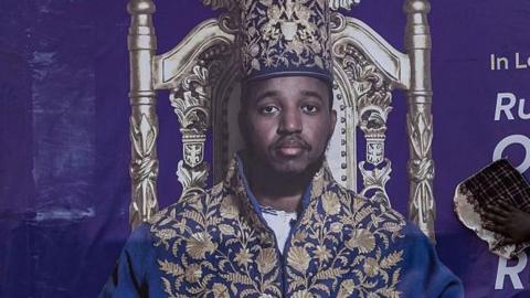 A portrait of King Oyo wearing an elaborate blue-and-gold ceremonial robe and matching crown sits on an ornate throne