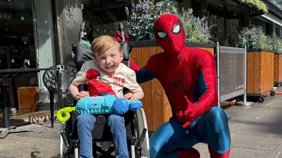 Photo: theukpulse.co.uk Noah Connell, who has short, blond hair, is in his wheelchair with a stunt man dressed as Spider-Man, complete with mask
