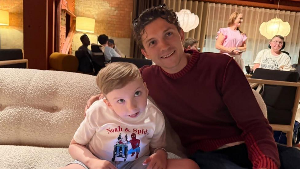 Noah Connell is sitting next to Tom Holland who has his arm round him. Noah has short, blond hair and is wearing a white T-shirt that says Noah and Spider-Man with a picture depicting him in his wheelchair with Spider-Man next to him. Holland has short, curly hair and is wearing a maroon jumper. They are both looking at the camera.
