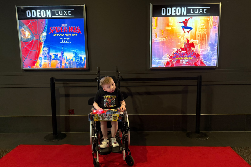 Noah Connell is sitting in his wheelchair on a red carpet with two Spider-Man cinema posters behind him. He has short, blond hair and is wearing a black Spider-Man T-shirt. He has a box containing a Spider-Man toy on his knee.