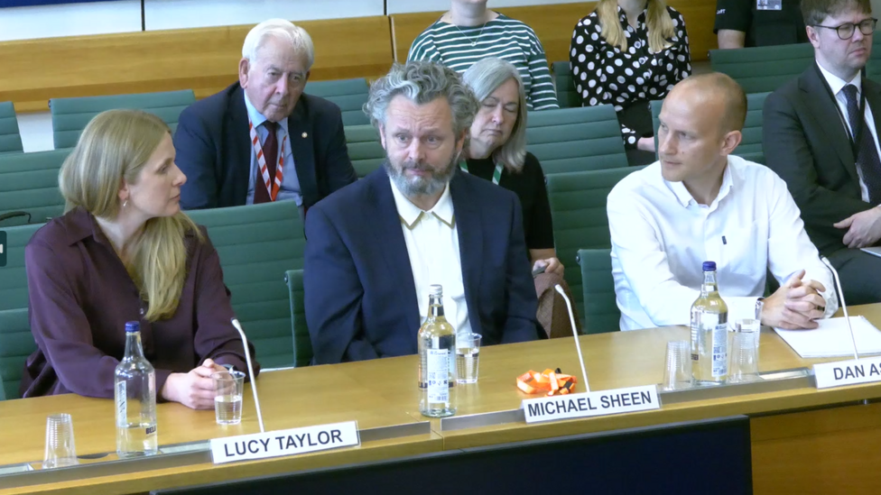 Michael Sheen in the centre in a dark blue suit and white shirt. On his right is a woman with long blonde hair and a purple top, in front of her is a sign that reads Lucy Taylor. On Sheen's left is a man with a white shirt, who is bald, in front of him is a sign that reads Dan.