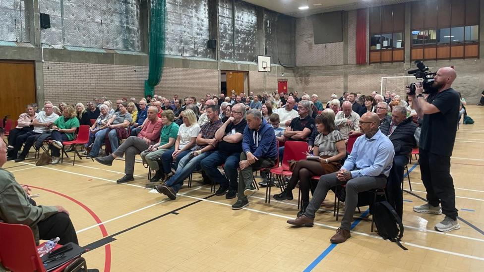 Around 160 people attend a public meeting at a leisure centre, with all sat in chairs in a sports hall.