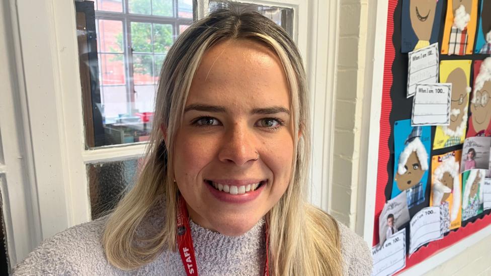 Sarah has long blonde hair and is wearing a grey jumper as she smiles at a camera in a school classroom setting.