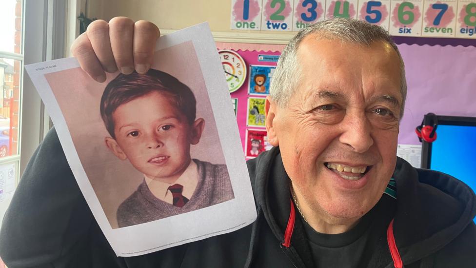 Colin has short grey hair and is wearing a black hoodie while he holds a picture of himself from school.