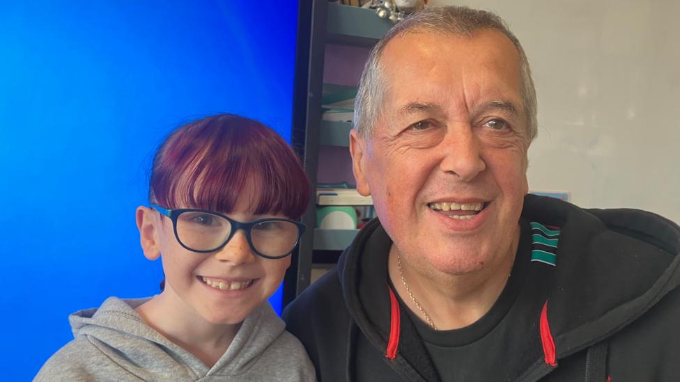 Poppy has her brown hair tied back in a ponytail and is wearing blue square glasses. Colin has short grey hair and is wearing a black hoodie.