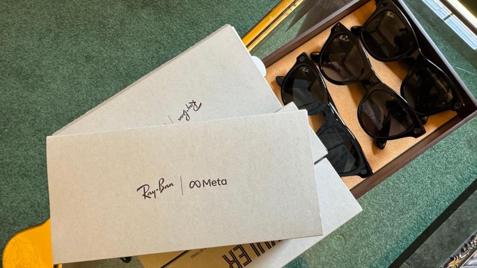 Four grey 'Meta Ray Bans' boxes stacked on top of each other, next to three pairs of sunglasses on a glass table.