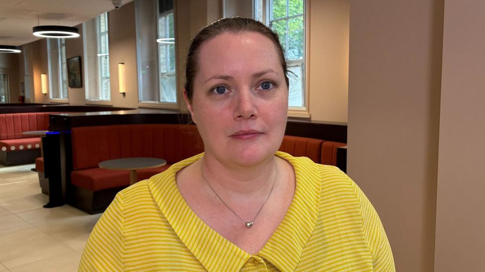A woman with brown hair, tied back, is looking at the camera. She is wearing a yellow shirt and a silver necklace.
