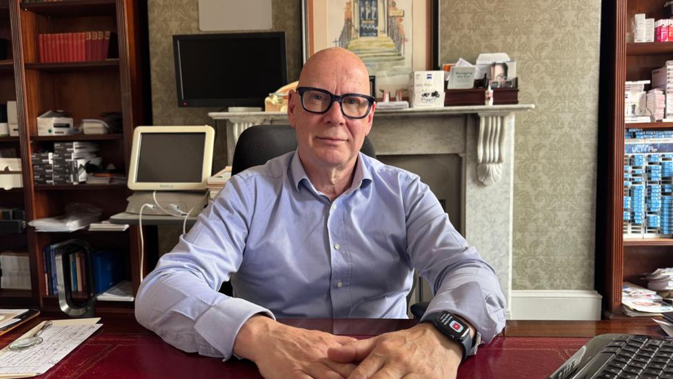A bald man wearing thick, dark glasses and a blue shirt is sitting at a desk in the office. His hands are on the desk and he is wearing a watch.