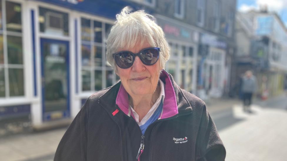 A woman wearing a fleece, sunglasses and a dark top with silver hair looks towards the camera while stood on a street, blurred in the background, with shop fronts visible