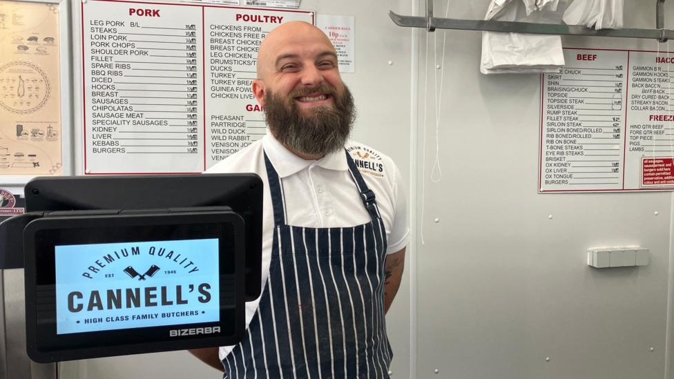 A man with a bushy beard smiles while stood wearing an apron and white shirt behind a butcher shop counter.
