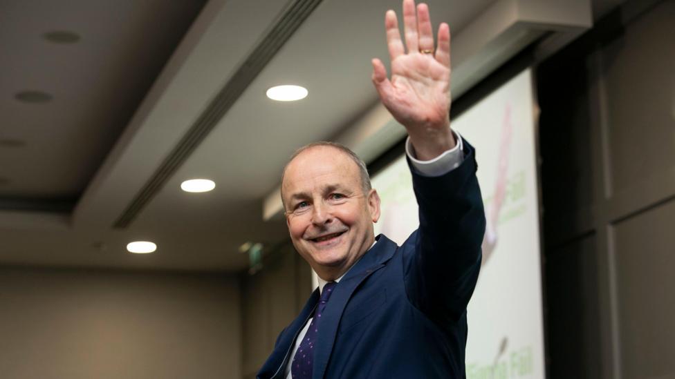 Micheál Martin, a man with short, greying hair, waves and smiles. He is wearing a navy suit, a white shirt and a navy and pink patterned tie. There is a screen on the wall behind him and ceiling spotlights above him.
