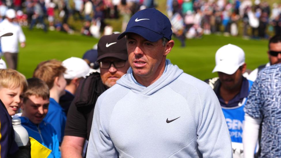 Rory McIlroy is wearing a light blue hooded sweatshirt and a navy blue baseball cap. Both items have the Nike logo on them. Behind him is a green golf course and hundreds of spectators.