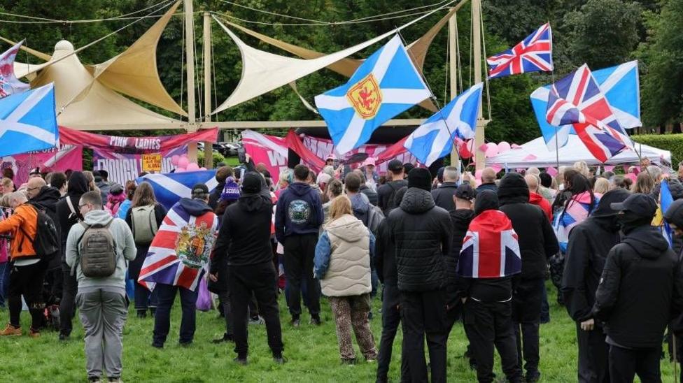 Crowd gathered in a park waving Scottish and Union flags.