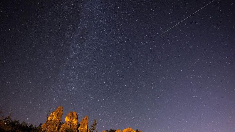 A rocky outcrop and the tops of some trees appear in the bottom left corner underneath a starry, indigo-coloured sky with a meteor streaking across in the top right corner.