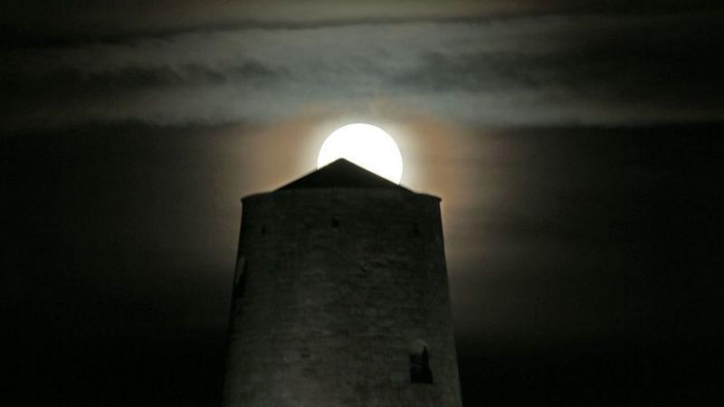 A bright full moon rises above a castle tower made of grey stone with a small peaked roof. There are some layers of cloud in the distance of a dark black sky.