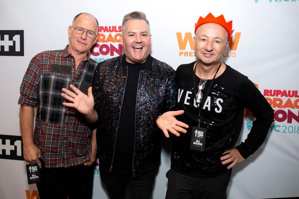 Three people standing in front of an event backdrop with visible logos and text including “RuPaul’s DragCon NYC 2018”, “WOW Presents Plus” and “#1”. The person in the centre wears a dark sequined jacket and raises both hands. The person on the left wears a plaid shirt and a black event pass on a lanyard. The person on the right wears a black sequined top with “VEGAS” printed on the front, a red paper crown, glasses hanging from a cord, and an “All Access” event pass.