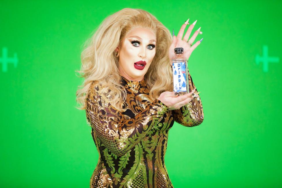 RuPaul's Drag Race UK - The Vivienne Clear plastic water bottle with a handwritten label reading “Drippin” and blue water-drop illustrations, held by Drag Queen The Vivienne on set of Drag Race. The Vivienne is wearing a black and gold patterned outfit and has long light-coloured hair, long pointed fingernails, and jewellery on several fingers.