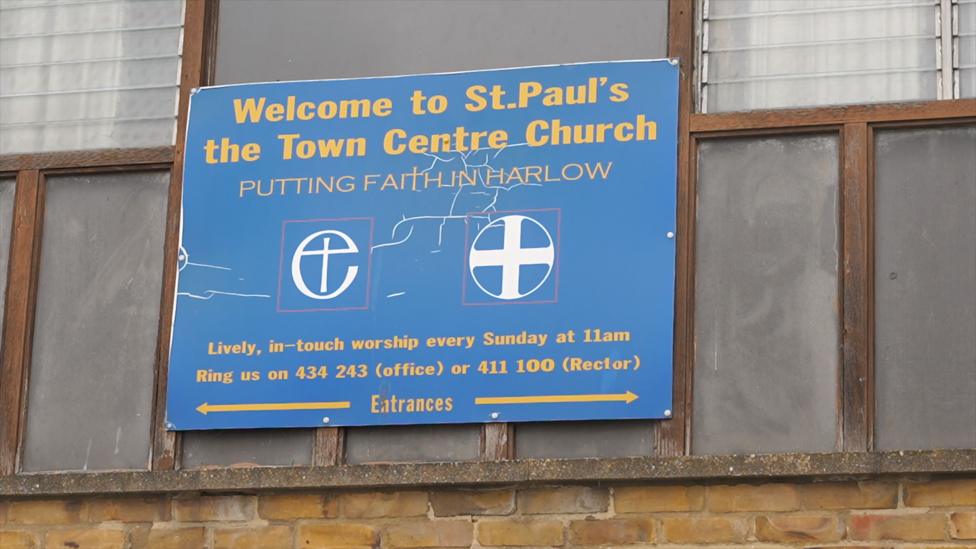 An old-looking sign attached to the side of a building. It is yellow text on a blue background. The sign says "Welcome to St Paul's the town centre church – putting faith in Harlow". There are also two logos based on crosses and contact details for the church.
