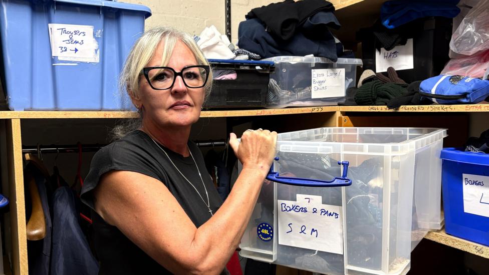 Kerrie Eastman looking down the lens, while pulling a box labelled "boxers and pants" off a shelf in a storeroom. She is standing in front of shelves that are holding other boxes, marked with labels including "trousers and jeans" and "hats". Kerrie has light blond hair, tied back, and she is wearing a short-sleeved black dress, with a long chain around her neck.