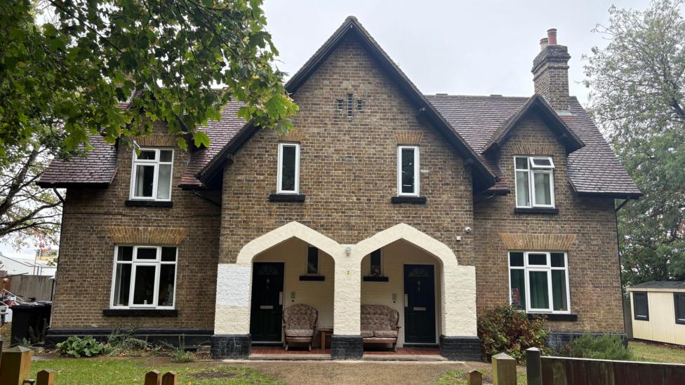 A semi-detached pair of Victorian cottages, set back from the road by a front garden. The image is taken front on, looking directly towards two archways that lead to each property's front door. Two arm chairs can be seen underneath the archways, in a covered area in front of the entranceways. Above each archway is a narrow, rectangular window and above that the apex of an imposing triangular roof line. On either side of the entrance and a little set-back are the side wings of the properties, with windows on both the ground and first floor. The image is framed by a mature-looking tree on the left and other trees are in the background on the right. There is a chimney in the roof on the right.