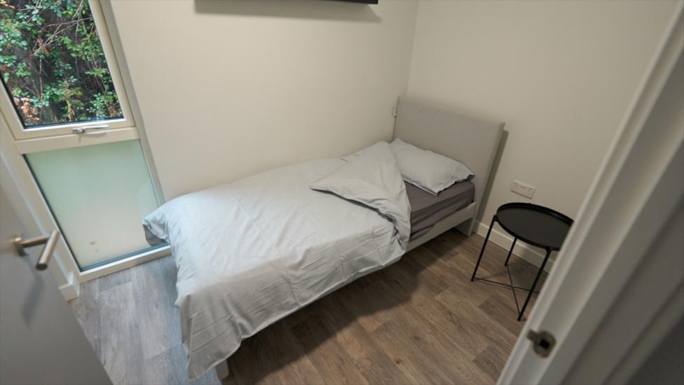 A single, made-up bed in a small bedroom, with white walls and wooden-effect flooring. There is a window at the foot of the bed, on the left of the image, looking out onto bushes. At the head-end of the bed is a low, circular black table.