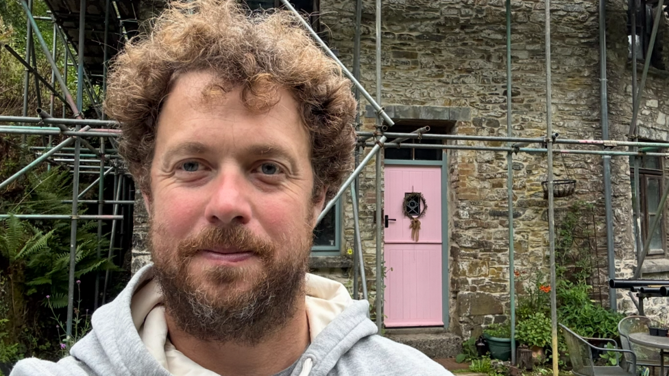 A person is standing in the foreground taking a selfie in front of a stone cottage that is covered in metal scaffolding.