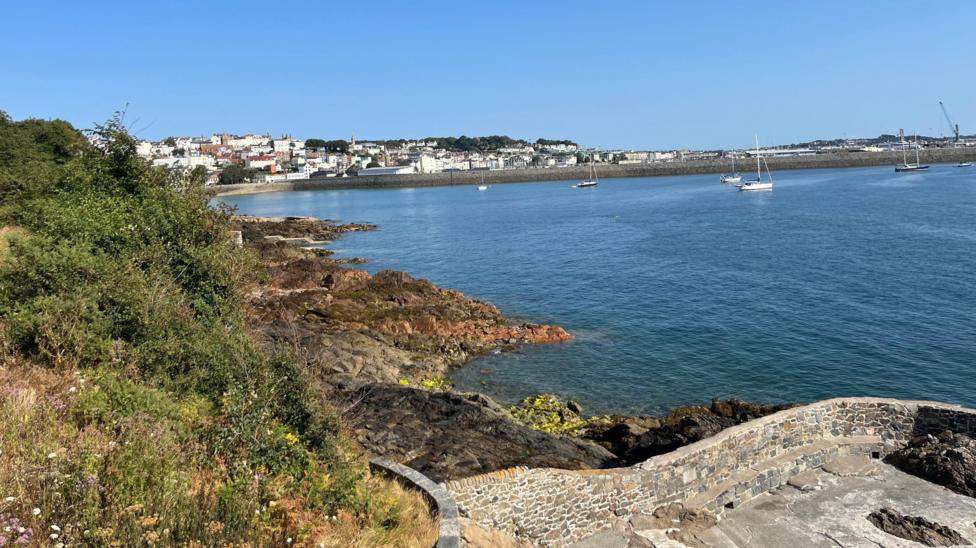A bright coastal harbour scene is shown under a clear blue sky, with calm blue water filling most of the image and several small sailing boats anchored offshore.