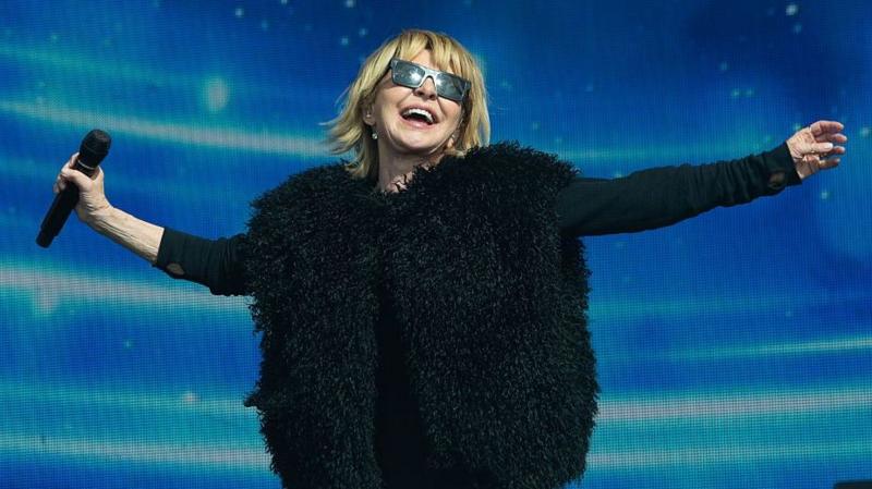 Lulu on stage during a performance. She has her arms outstretched and is smiling towards the sky. She is wearing dark sunglasses, a black top and a fluffy black jacket with no arms over the top.