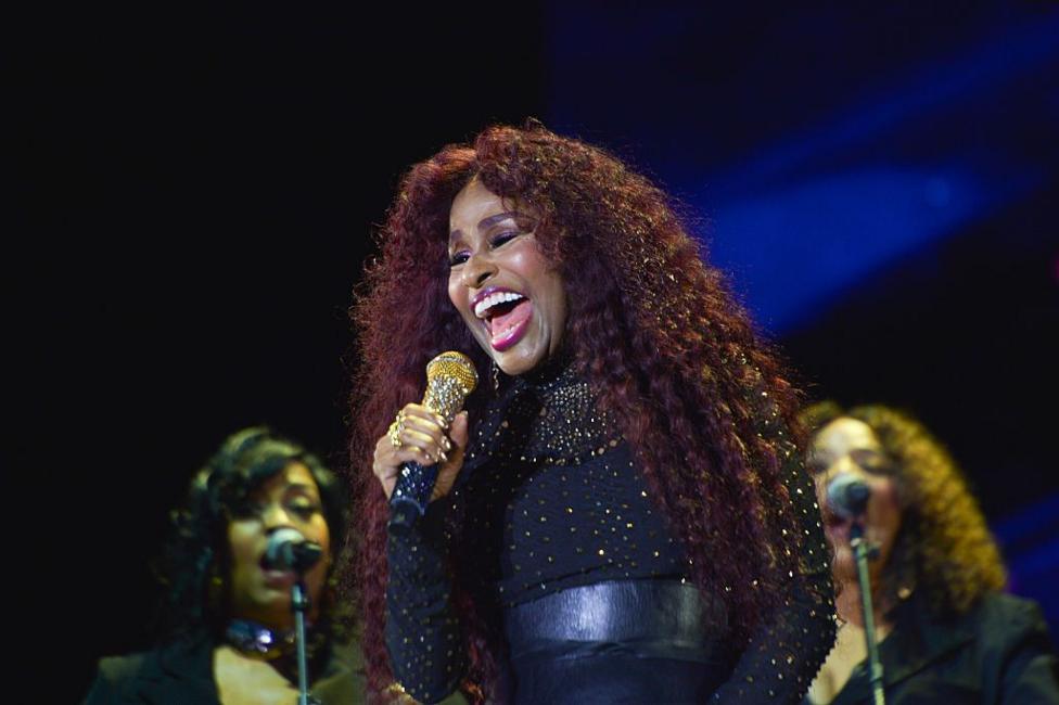 Chaka Khan singing into a gold mic on stage with two backing singers behind her. She has long, brown hair and is wearing a black, sparkly top