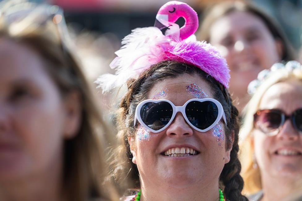 Woman at a festival. She is wearing white-rimmed, heart-shaped sunglasses, and a hat shaped like a pink flamingo. She has multi-coloured glitter on her face.