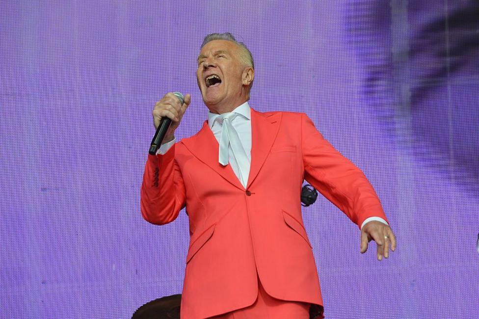 ABC's Martin Fry singing into a microphone on stage. He is wearing a red suit, a white shirt and white tie.