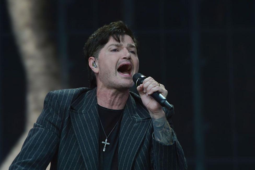 Danny O'Donoghue from The Script singing into a microphone on stage. He is wearing a black pin-striped jacket with a black top underneath. He has a large, silver cross around his neck.