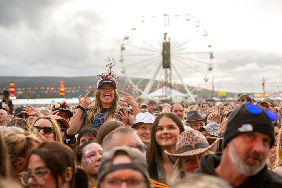 A crowd of people at a festival. One woman is sitting on someone's shoulders with lots of other people around her. She has an elaborate hair band over the top of a black baseball cap and is sticking her tongue out while gesturing with her hands