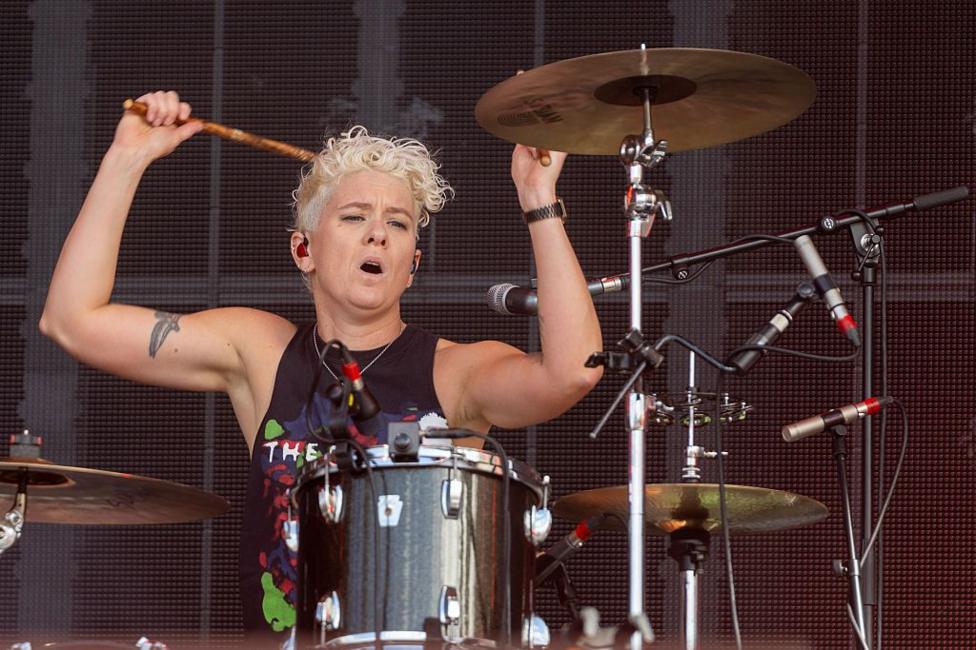 Texas drummer Cat Myers of playing on stage. She has short, curly, blonde hair and is wearing a black vest. Her arms are raised with drumsticks in her hands about to strike the drums